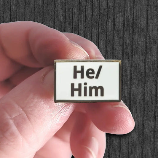 Hand holding a badge with 'He/ Him' pronoun text against a dark background