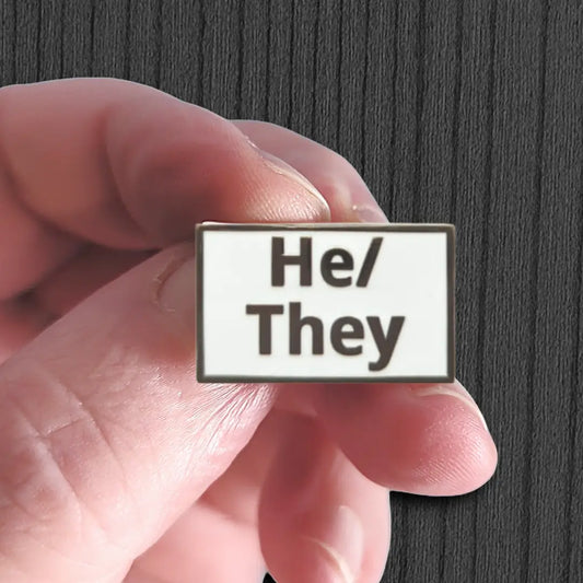 Pin with 'He/They' text held between fingers against a dark background
