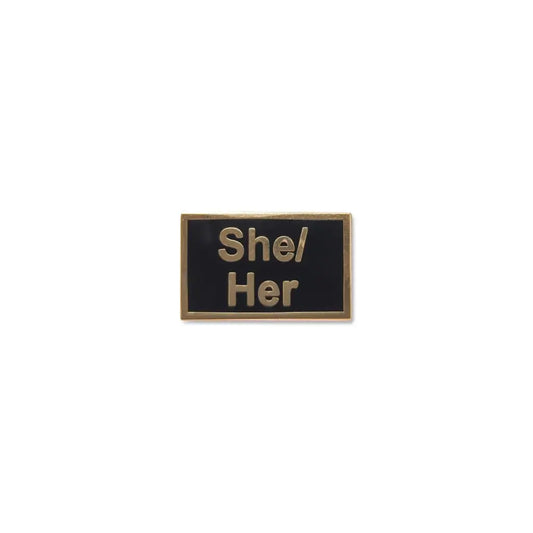 Elegant black She/They lapel pin with raised silver-tone lettering and a metallic frame, offering a minimalist and durable pronoun display for daily wear or professional settings.