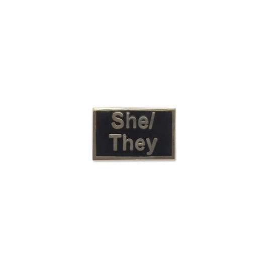 Elegant black She/They lapel pin with raised silver-tone lettering and a metallic frame.