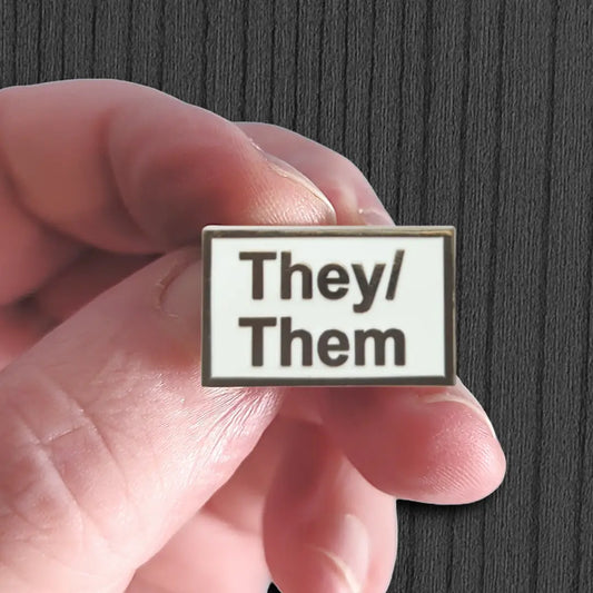 Hand holding a badge with 'They/Them' text against a dark background