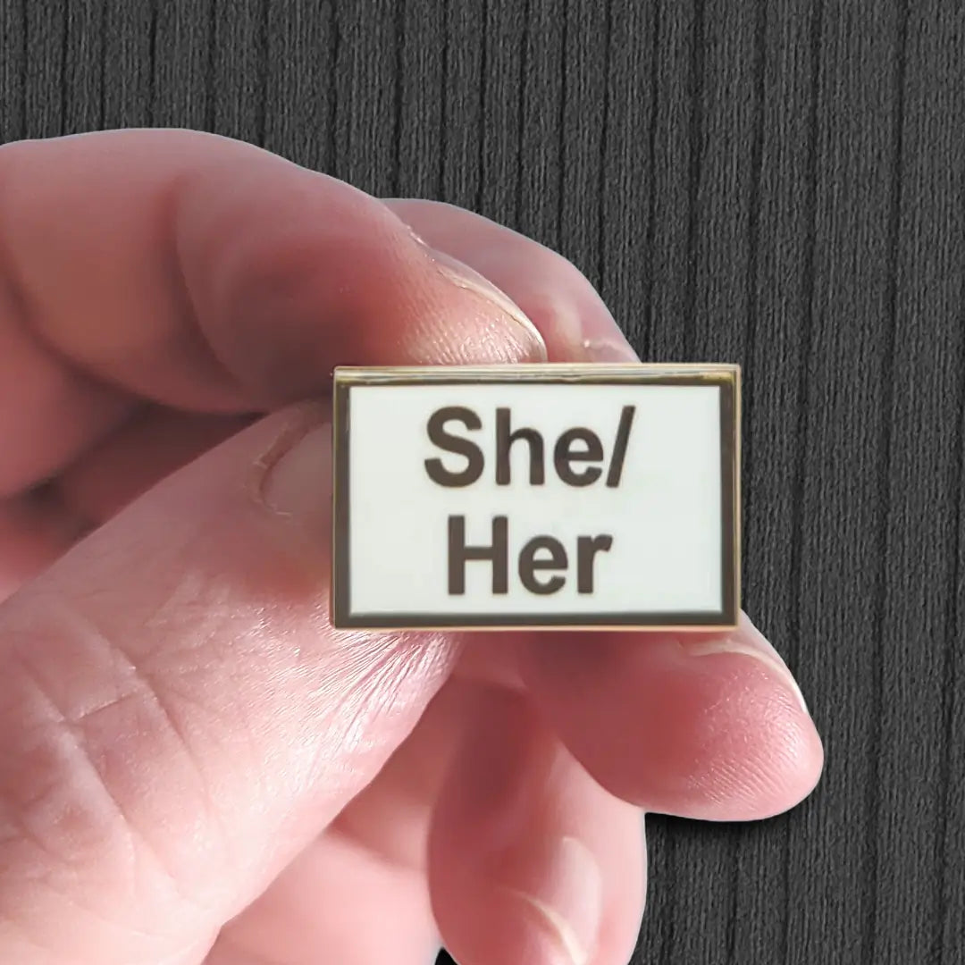 Hand holding a badge with 'She/Her' pronouns against a dark background