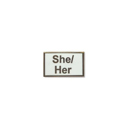 Rectangular She/Her lapel pin with a crisp white enamel background and bold black lettering, framed in a fine metallic outline a minimalist, gender-affirming accessory.