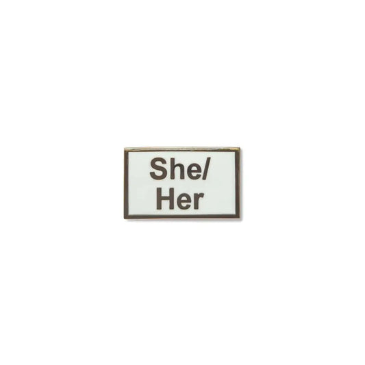 Rectangular She/Her lapel pin with a crisp white enamel background and bold black lettering, framed in a fine metallic outline a minimalist, gender-affirming accessory.