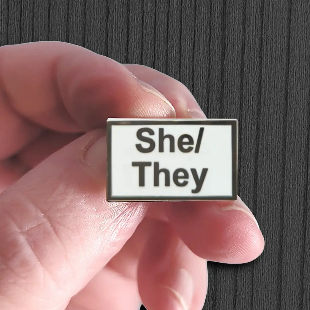 Hand holding a badge with 'She/They' pronouns against a dark background