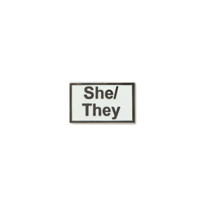 Rectangular She/They lapel pin with a white enamel background, bold black text.