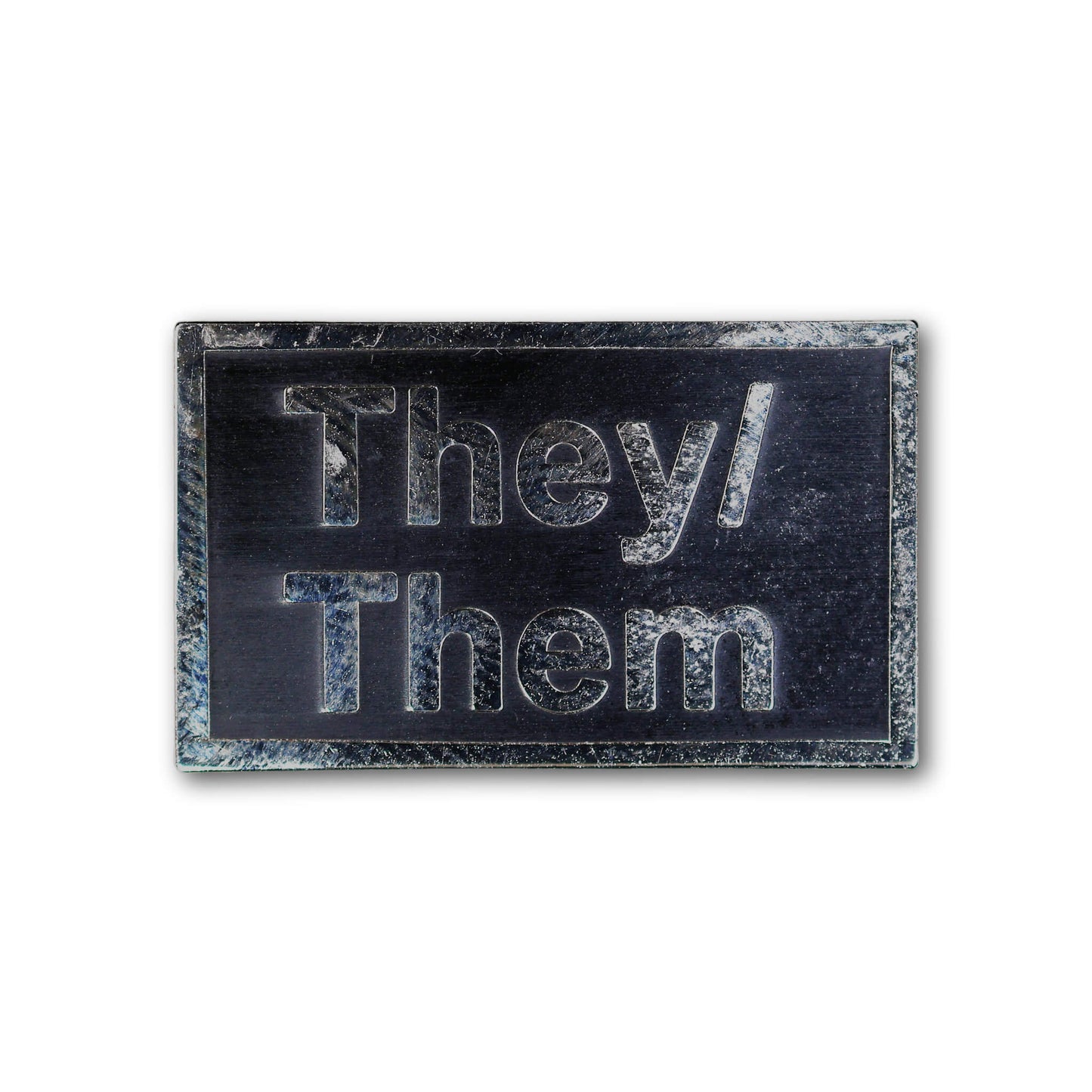 Rectangular metal badge with 'They/Them' engraving on a white background
