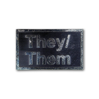 Rectangular metal badge with 'They/Them' engraving on a white background