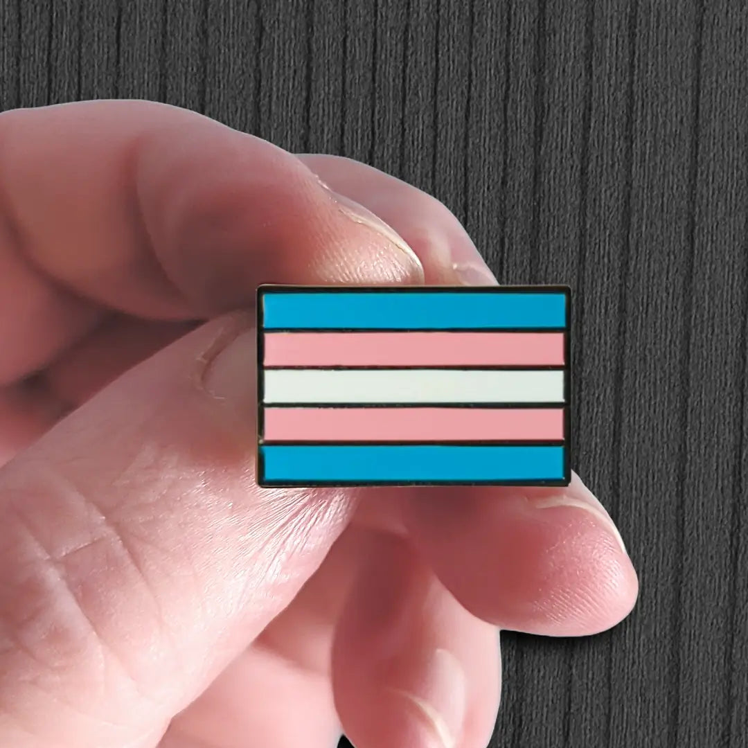 Transgender pride flag pin held between fingers against a dark background