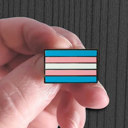 Transgender pride flag pin held between fingers against a dark background